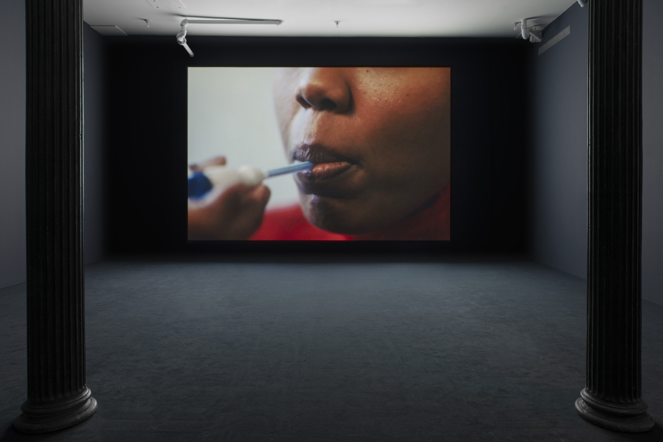 Carolyn Lazard: Two - way . Installation view ( Vital , 2025), Artists Space, 2025. Photo: Greg Carideo. Image CourtA darkened gallery space with dark blue walls and dark blue carpeting. A film is projected onto the farthest wall. The projection depicts a close - up of the lower half of a black person’s face. A hand is shown taking the person’s temperature orally with a thermometer.