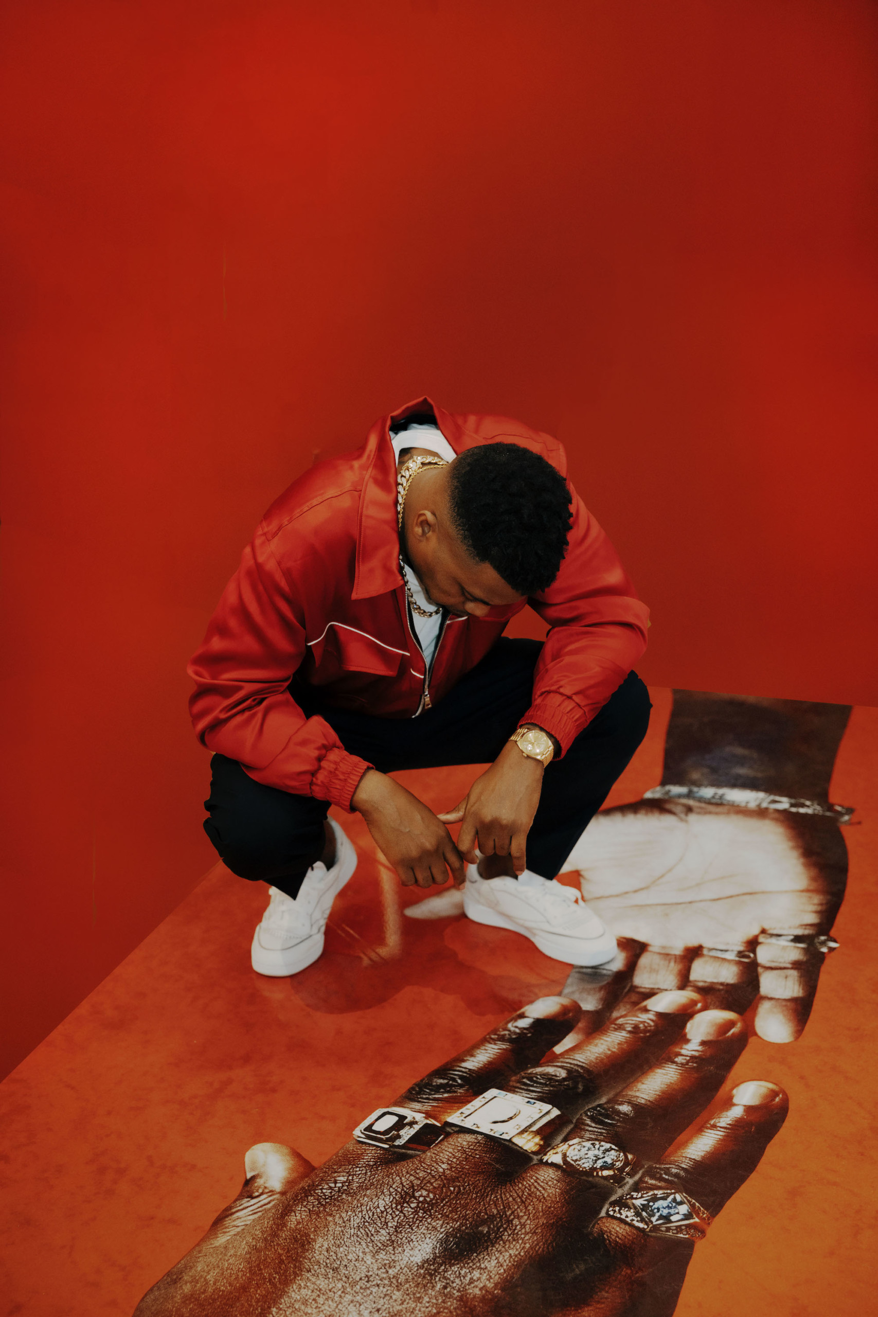 Man in red leather jacket against red wall squatting and looking at art of a hand on floor