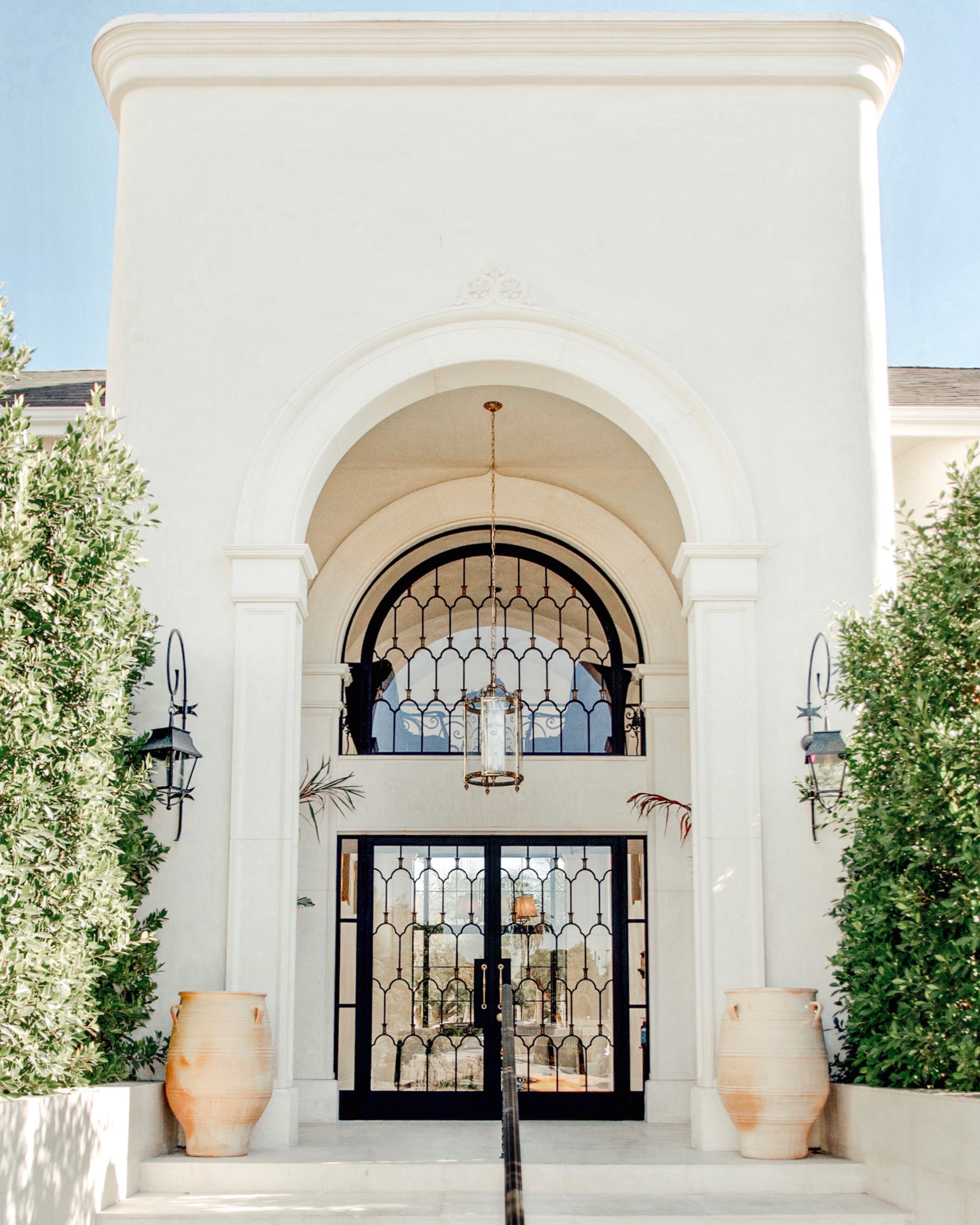 rustic hotel entrance flanked by tall trees and pottery, with large doors and mediterranean style chandelier
