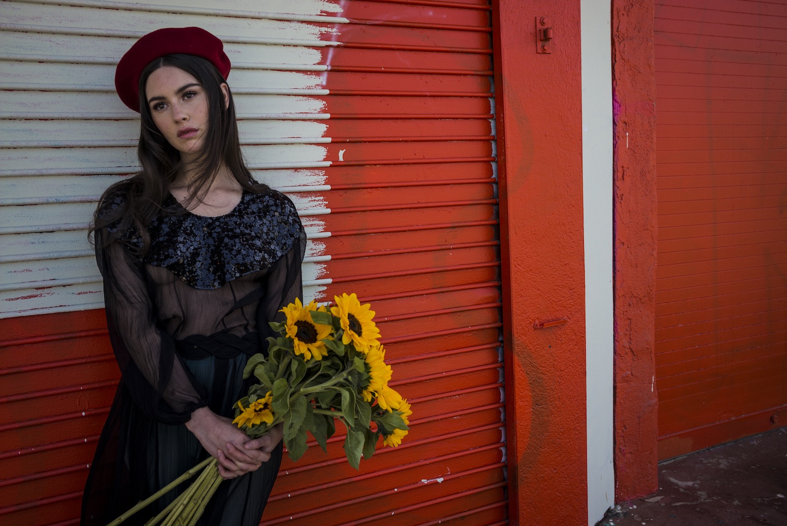 artists on tour gracie abrams against red wall wearing red beret and holding bouquet of sunflowers